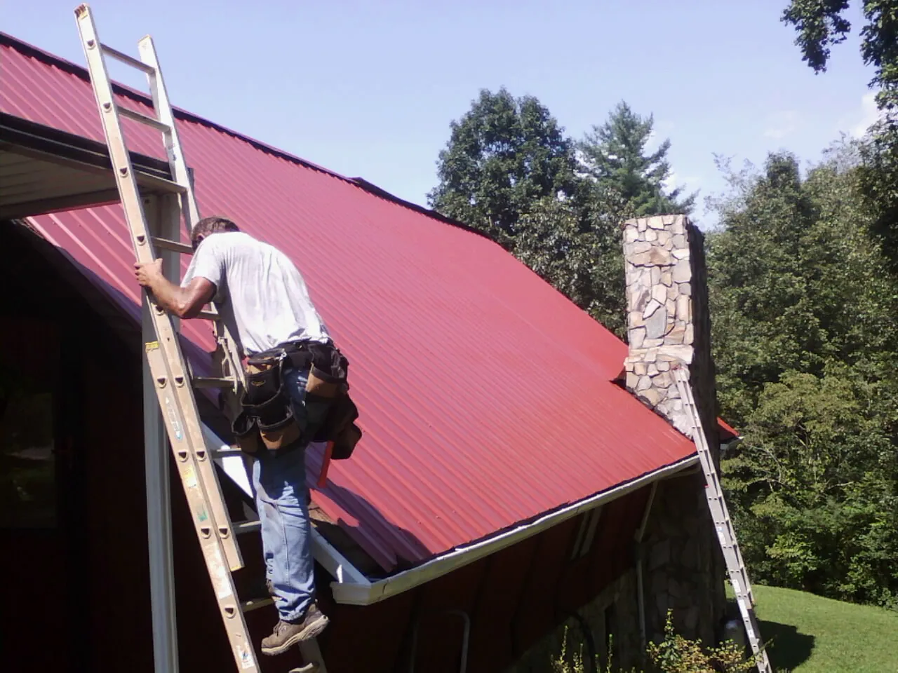 Roofer on a ladder beside a red metal roof and stone chimney — Soffit & Fascia Repair by Roofing Brothers in Centre Hall